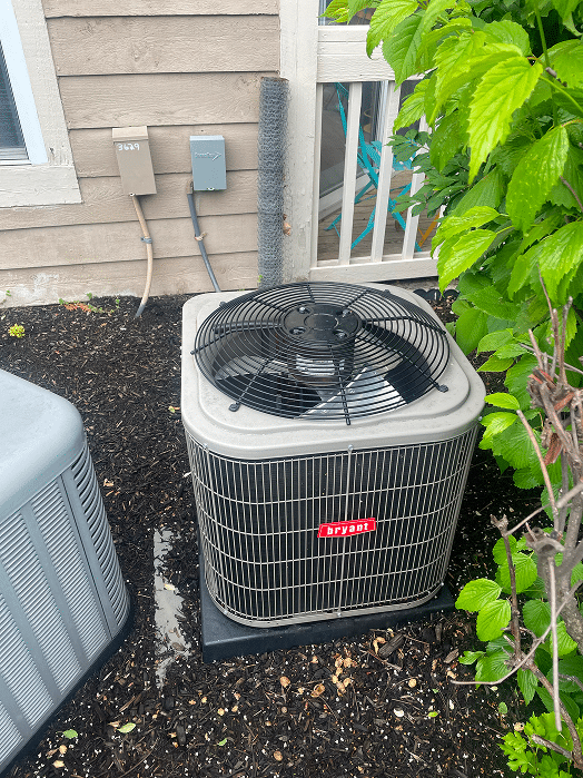 A Bryant brand central air conditioning condenser unit installed on a black pad in a mulched garden bed next to a residential home in Hilliard, Ohio.